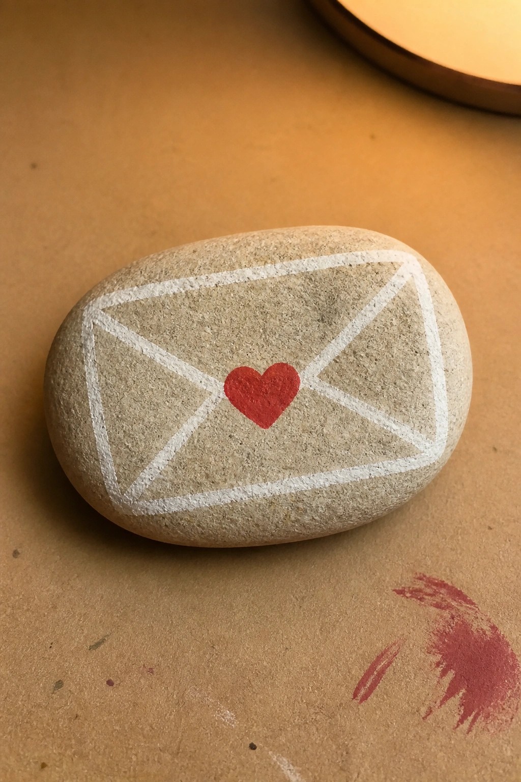 Rounded stone painted with white envelope outline and red heart on brown surface