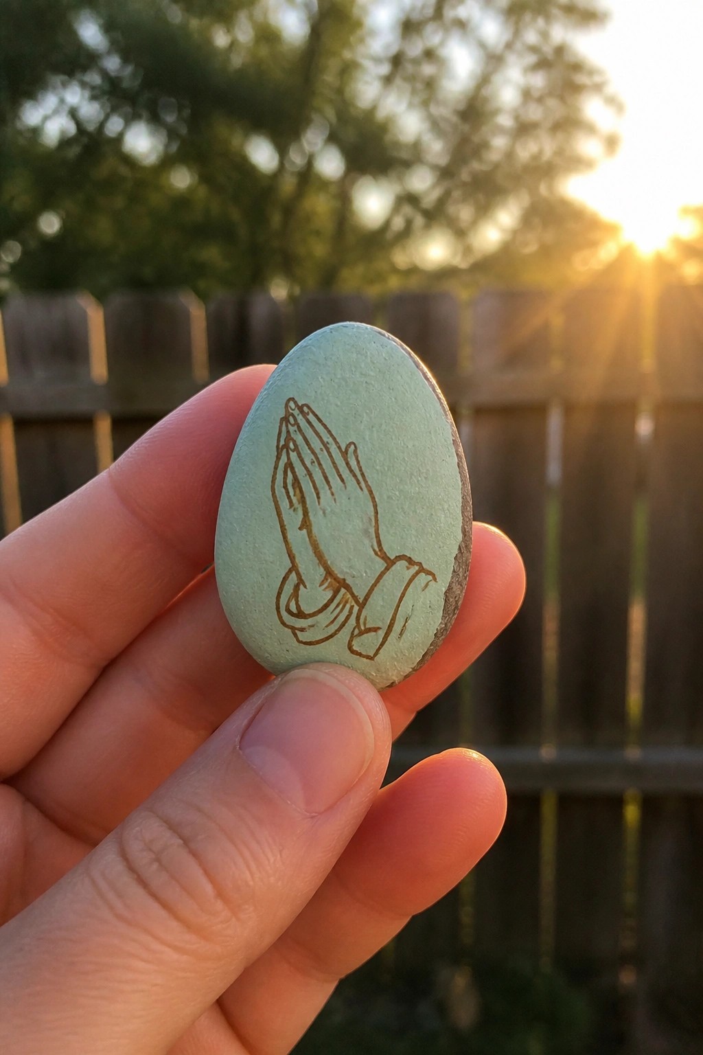 Hand holding light green stone engraved with praying hands at sunset