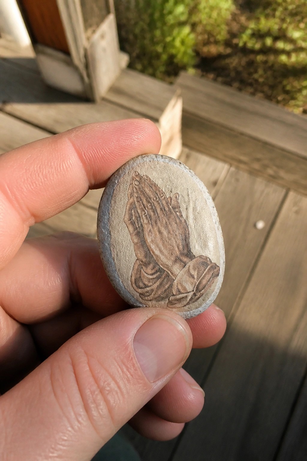 Hand holding oval stone engraved with praying hands on wooden deck