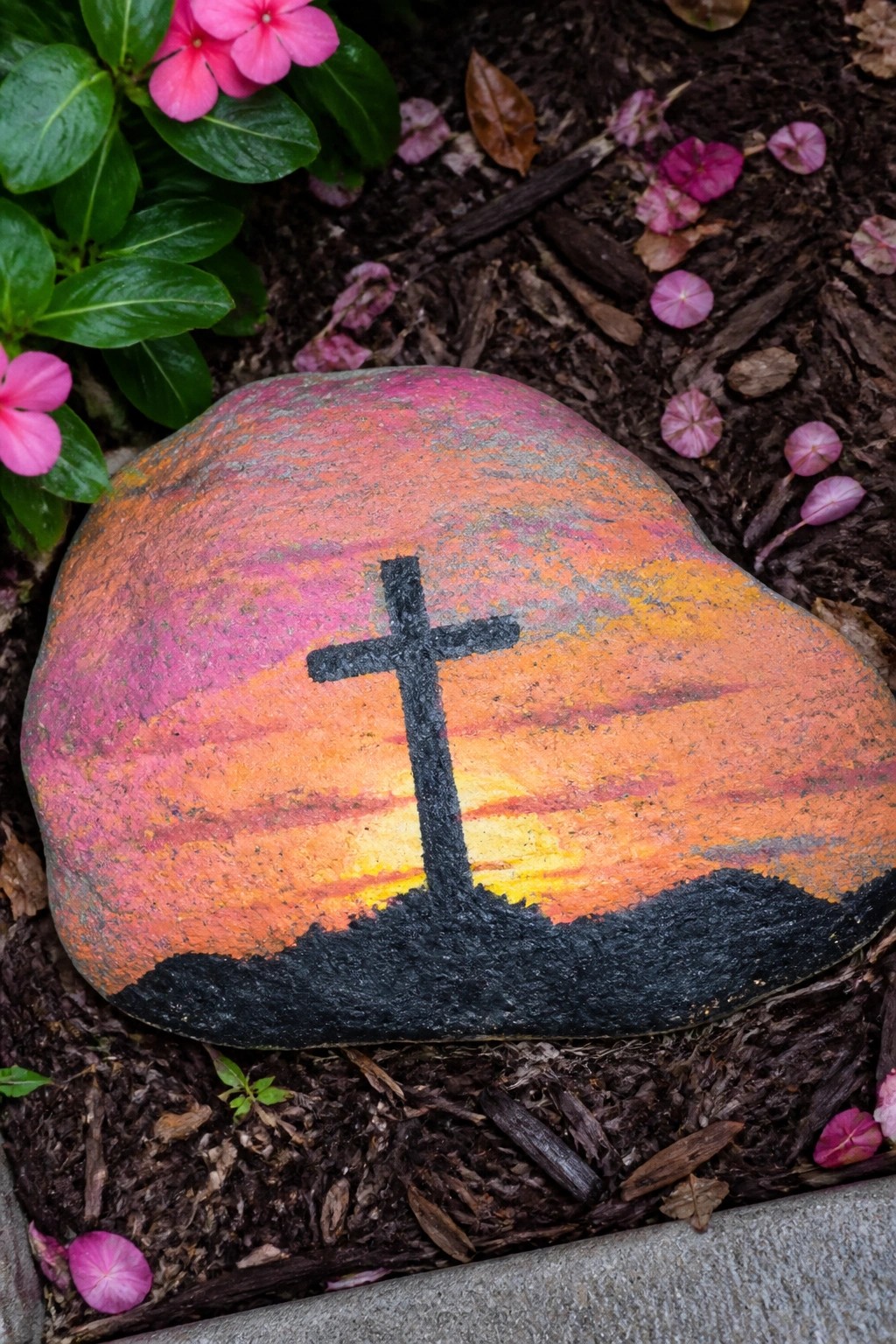 Painted rock with colorful sunset stripes and black cross amid pink flowers and mulch