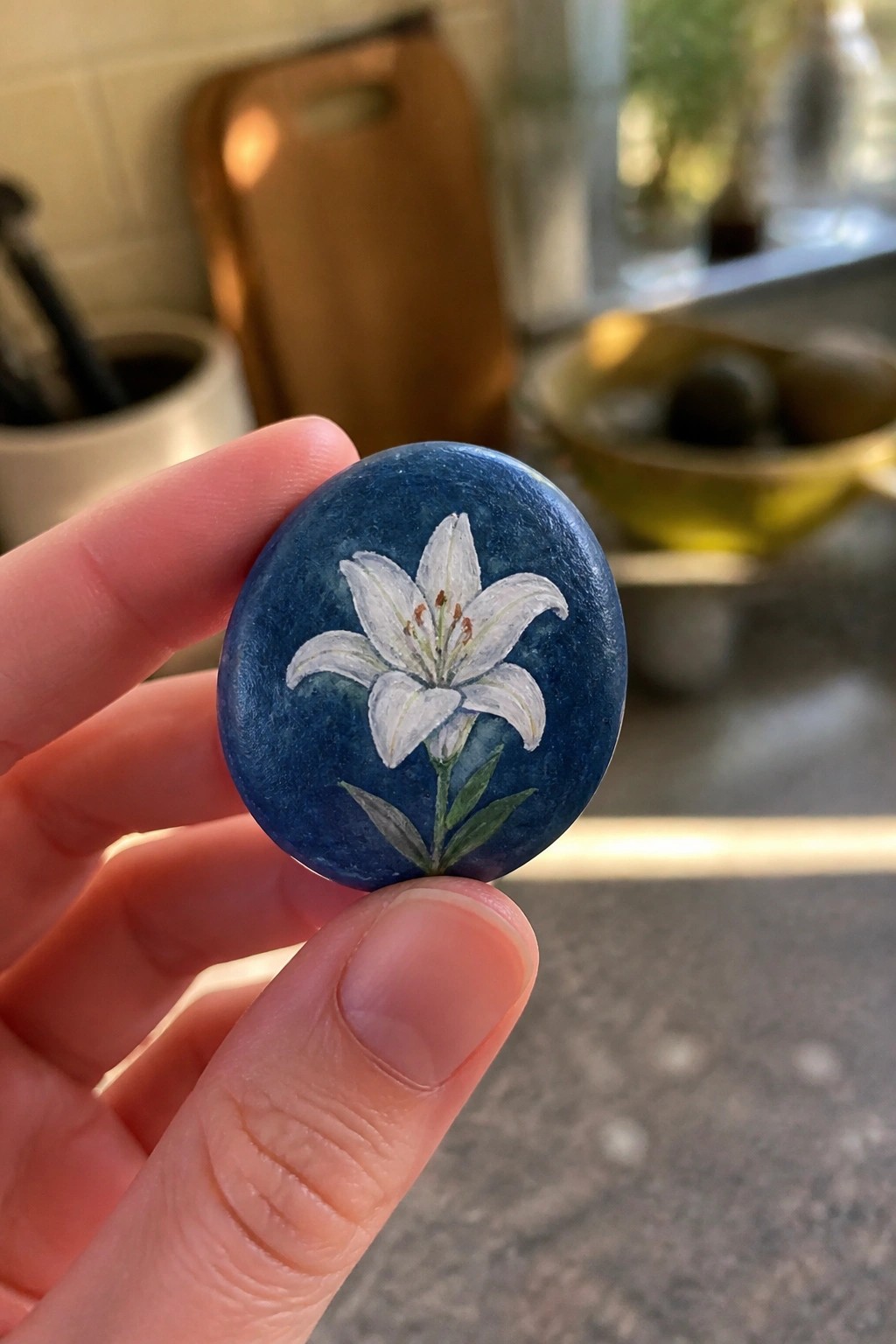 Hand holds blue oval stone painted with white lily and green leaves.