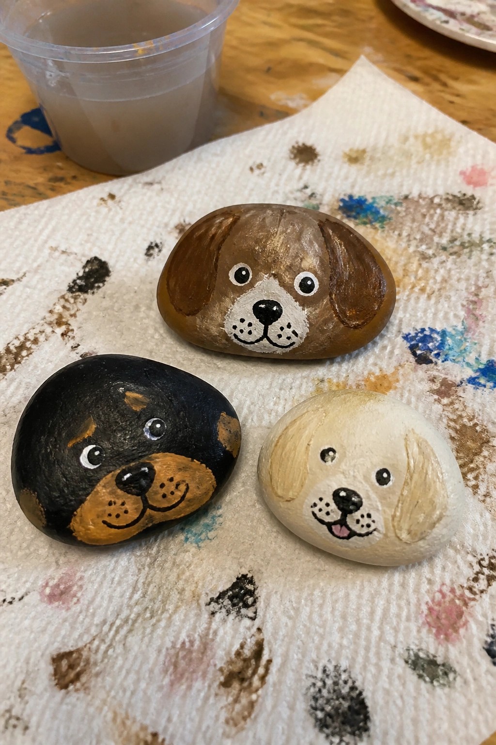 Three painted dog rocks on a splattered paper towel beside a plastic cup