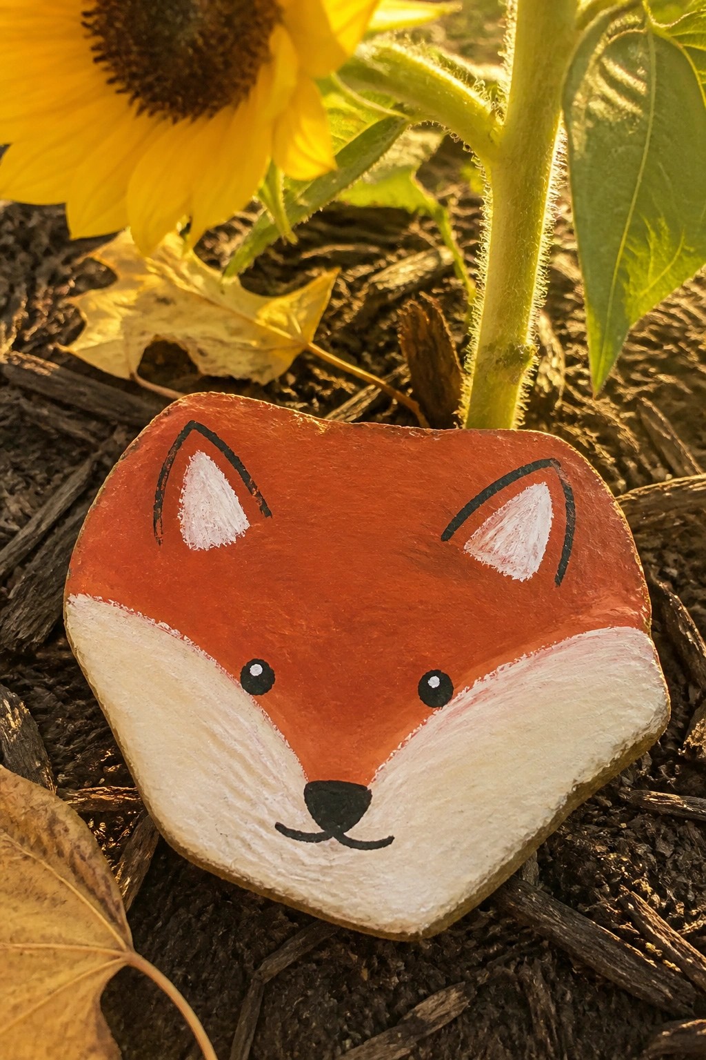 Painted orange fox rock with white muzzle and ears on soil near sunflower.