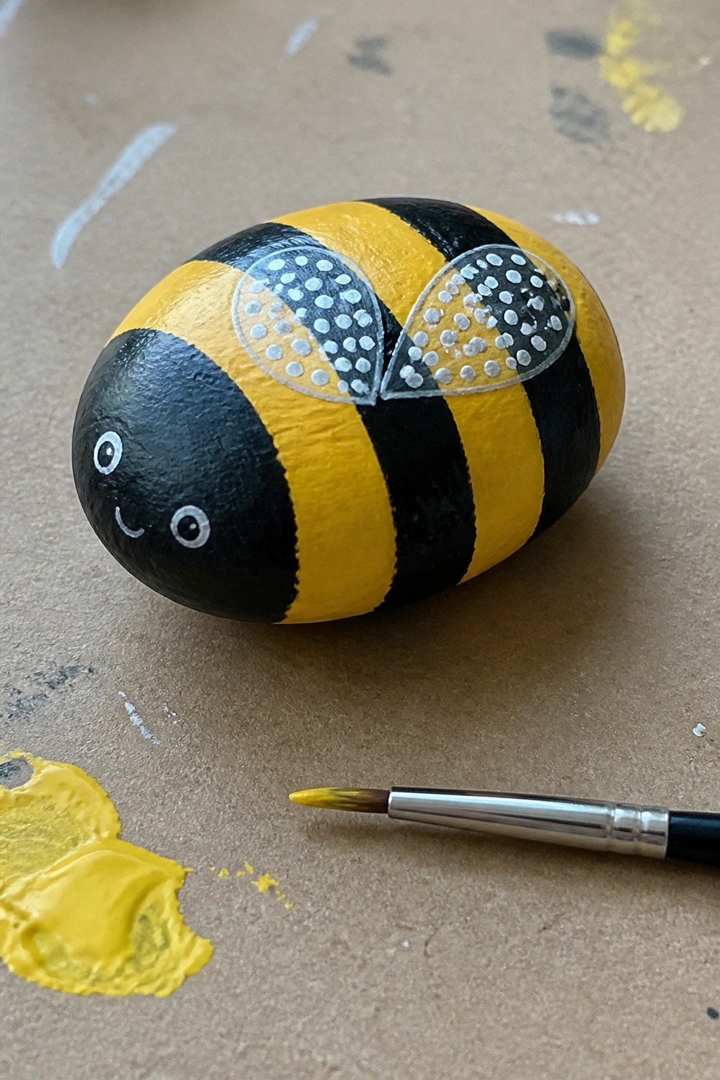 Painted bumblebee rock with yellow-black stripes, dotted wings, smiling face, and nearby paintbrush.