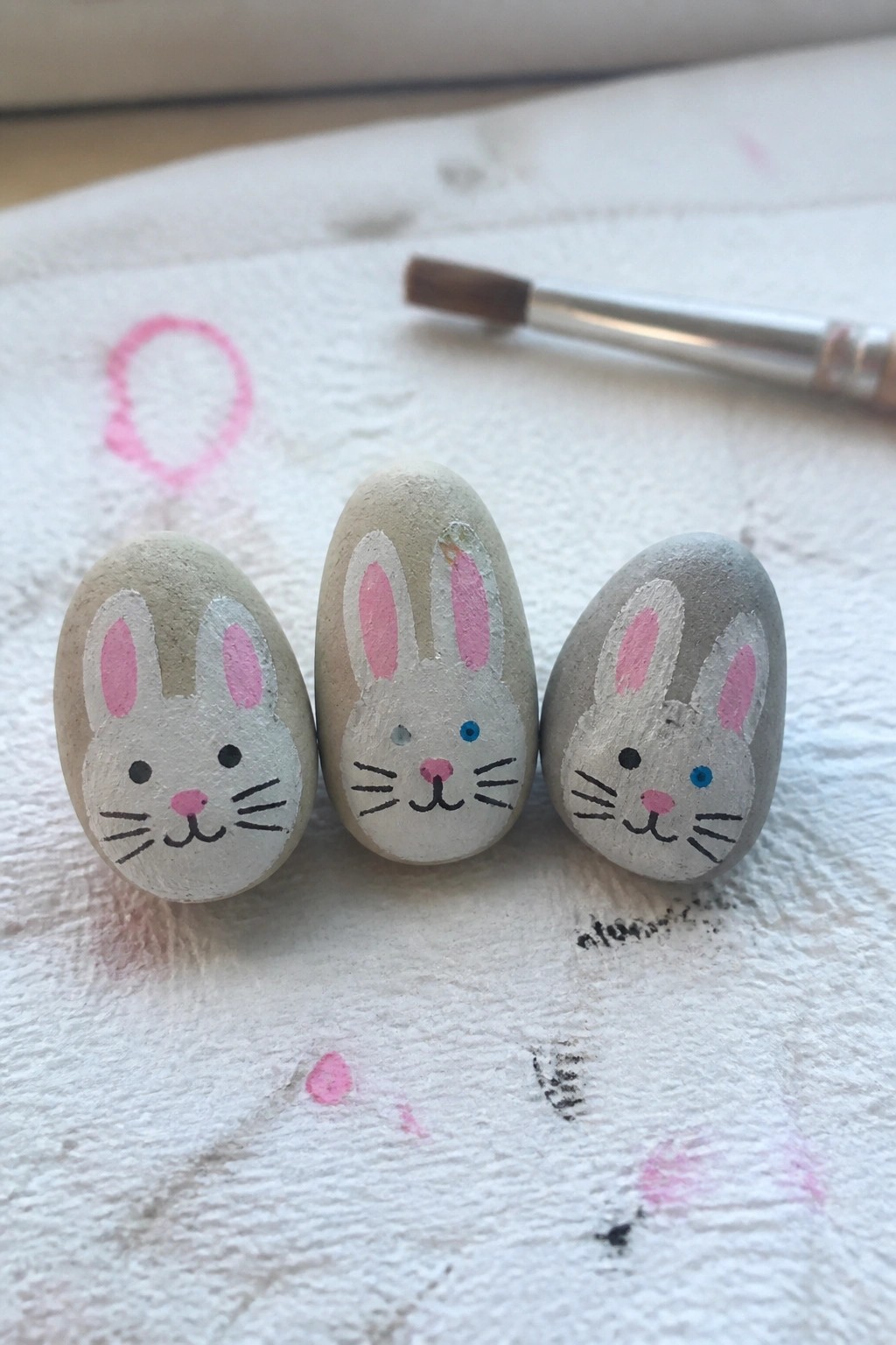 Three painted rocks as bunnies with pink ears beside paintbrush on white paper towel