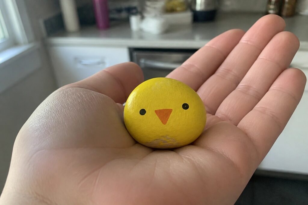 Hand holding smooth yellow rock painted as chick with black eyes and orange beak