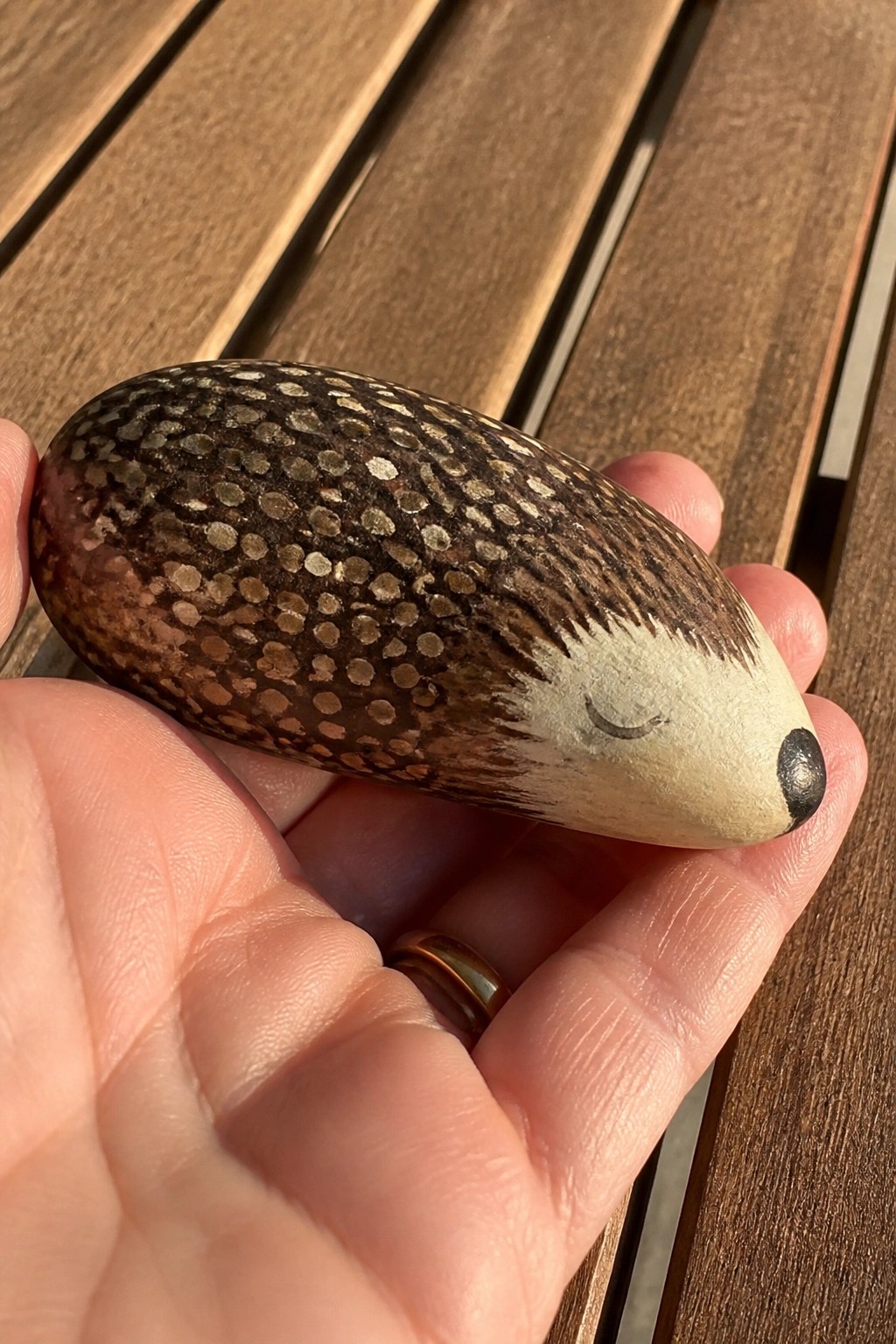 Hand holding hedgehog-painted rock with brown spots, white face on wooden slats.