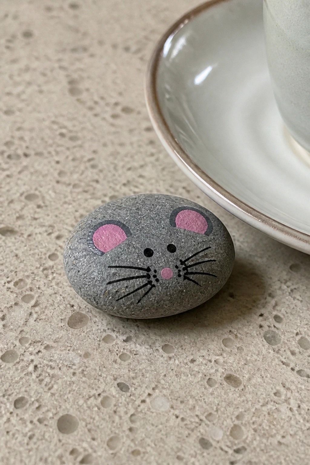 A small gray stone painted with a mouse face featuring pink ears, black eyes and whiskers, and a pink nose sits on a speckled surface.