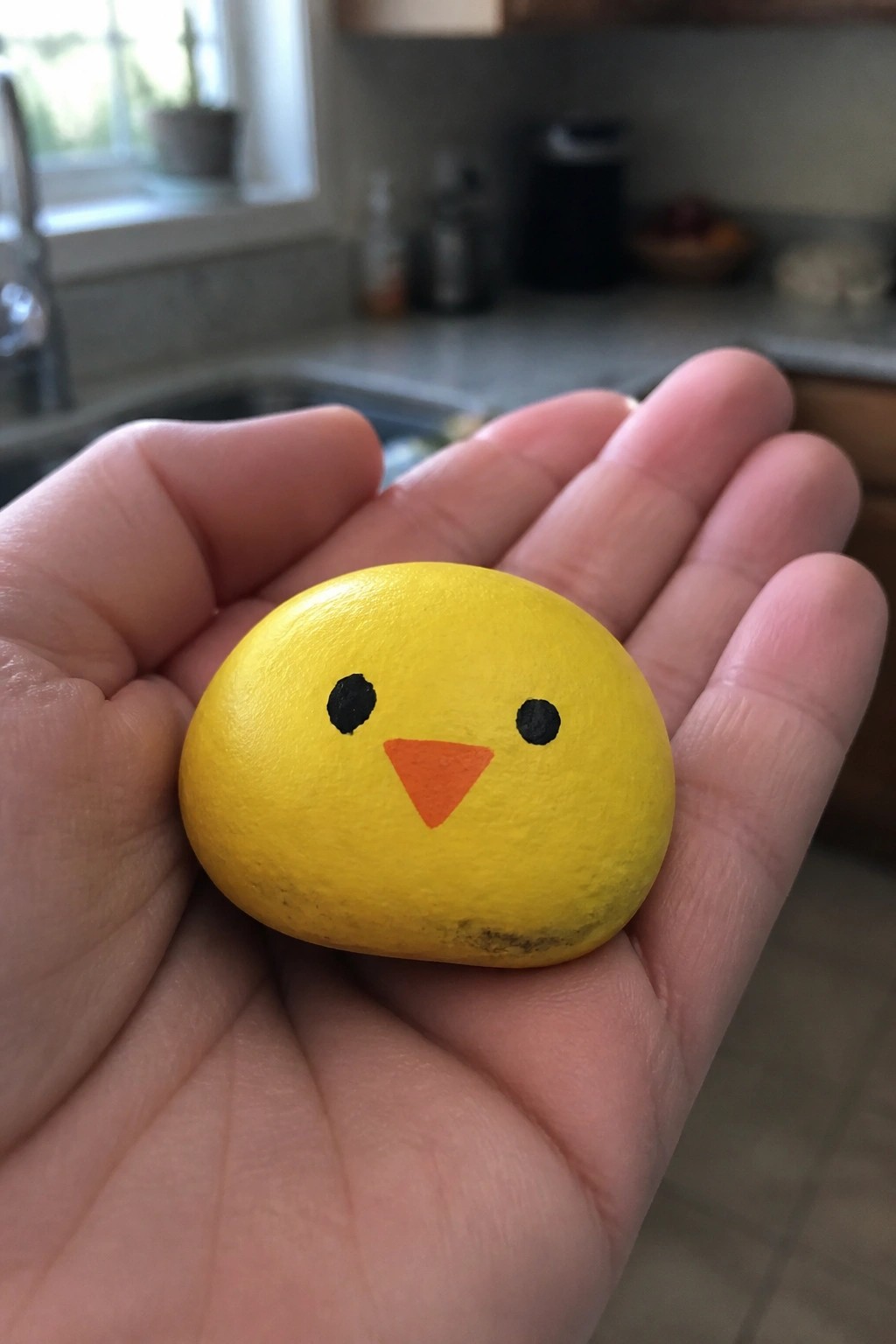 Hand holding smooth yellow rock painted as chick with black eyes and orange beak