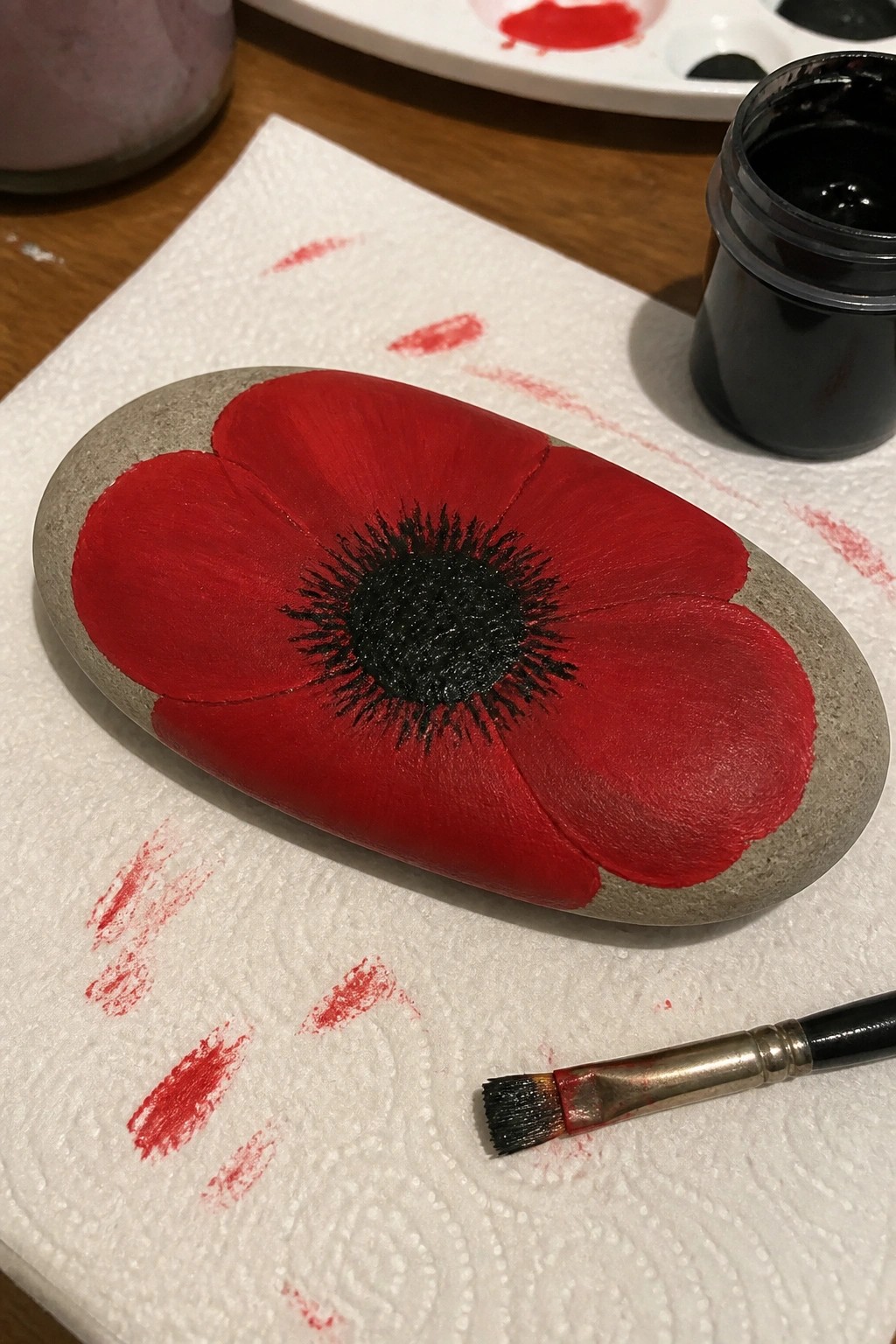 Gray oval stone painted with red poppy and black center beside paintbrush on towel.