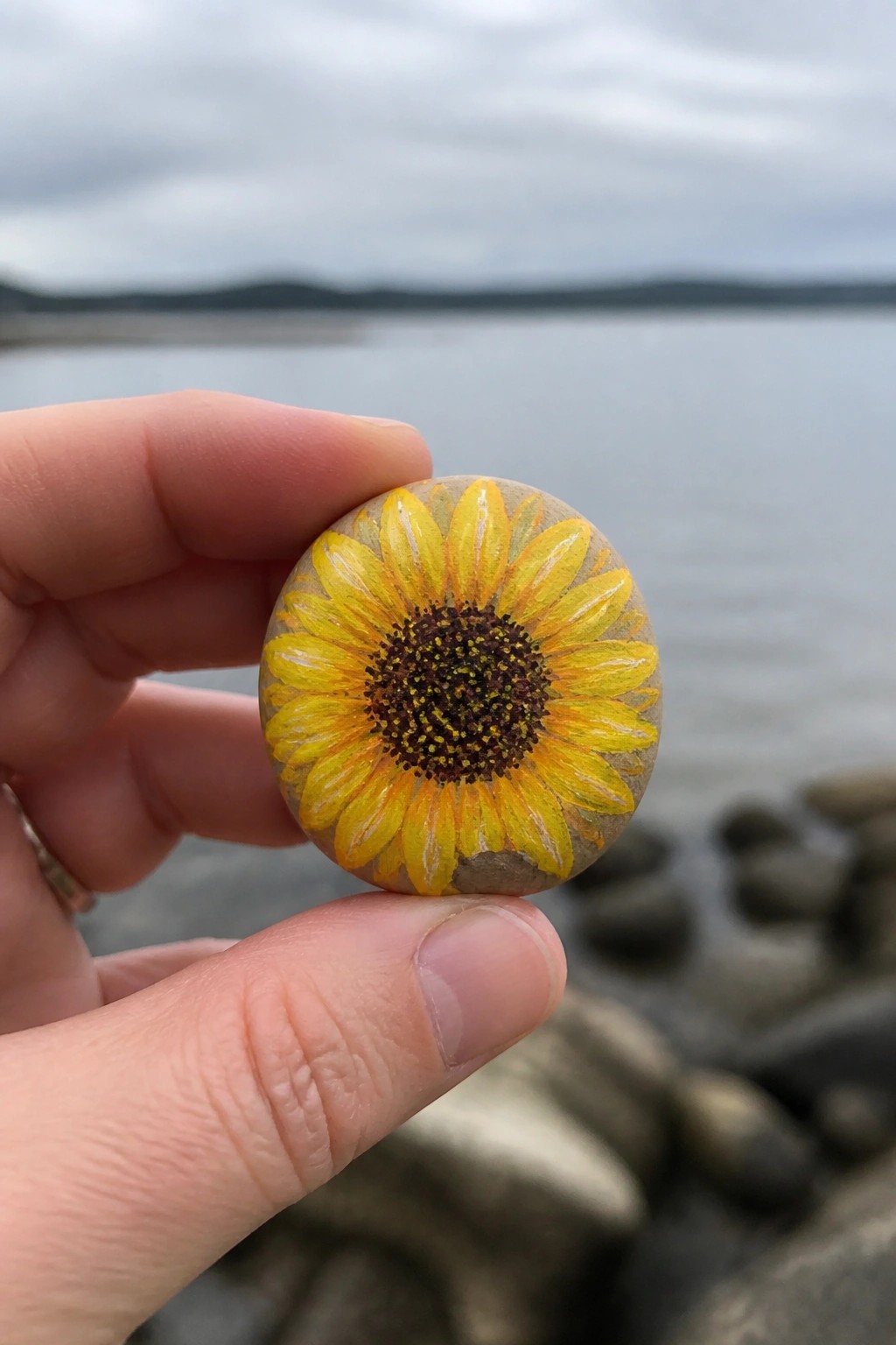 Hand holding round stone painted with bright yellow sunflower by water.