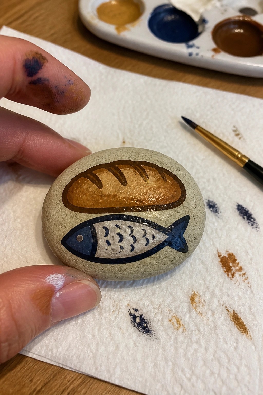 A hand holds a smooth gray stone painted with a brown loaf of bread above a blue and white fish on a paper towel next to paint supplies.