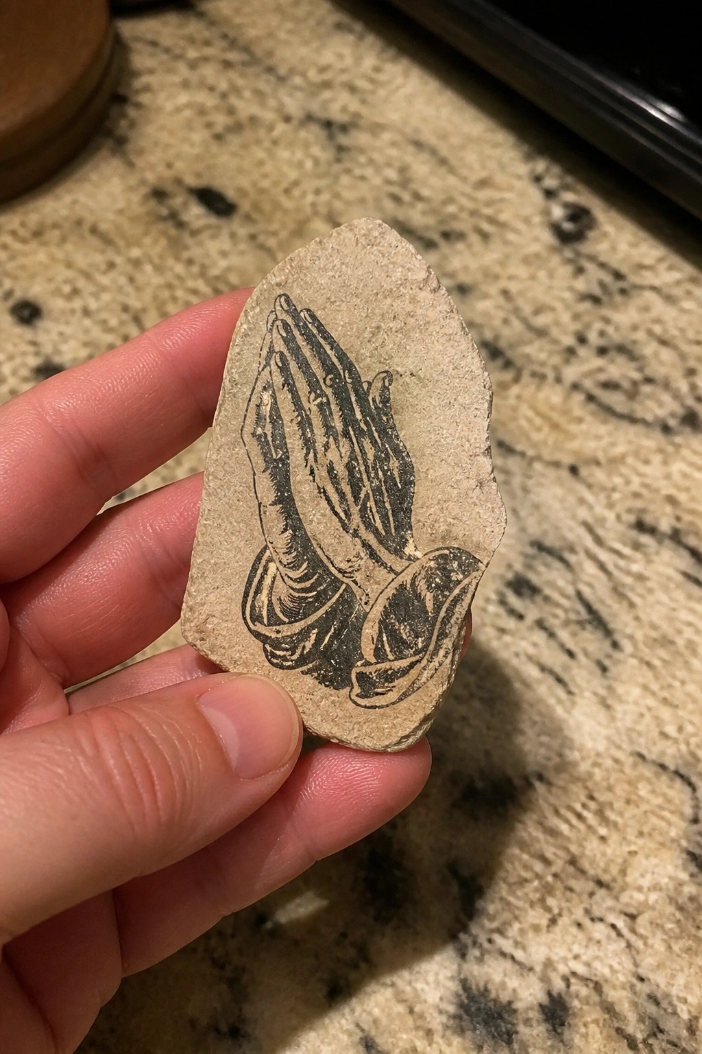 Hand holding irregular beige stone printed with black praying hands on granite counter