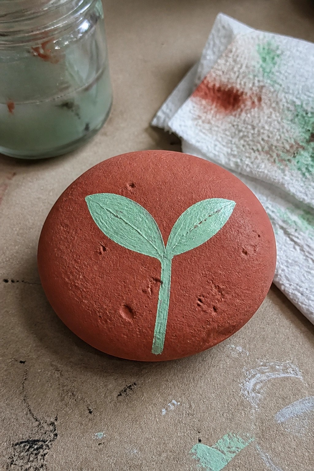 Red painted rock with green sprout beside glass jar and stained paper towel