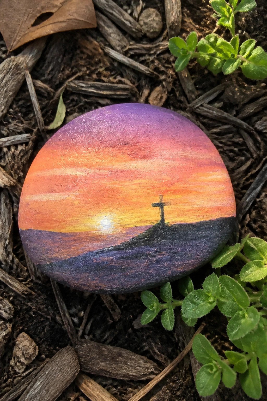 Painted rock with vibrant purple-orange sunset, glowing sun, and silhouetted cross on hill.