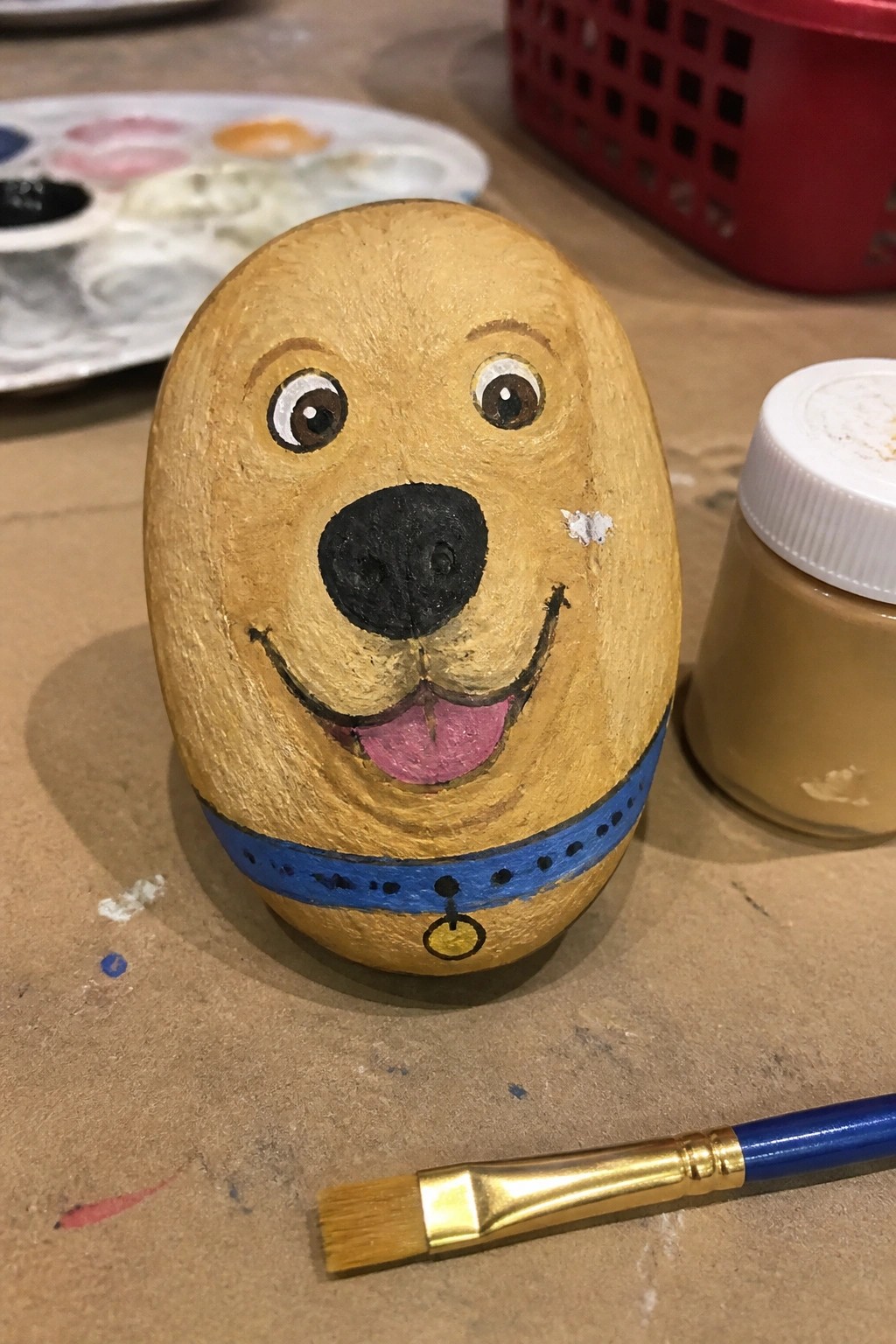 Painted golden dog rock with blue collar beside paintbrush and tan paint jar