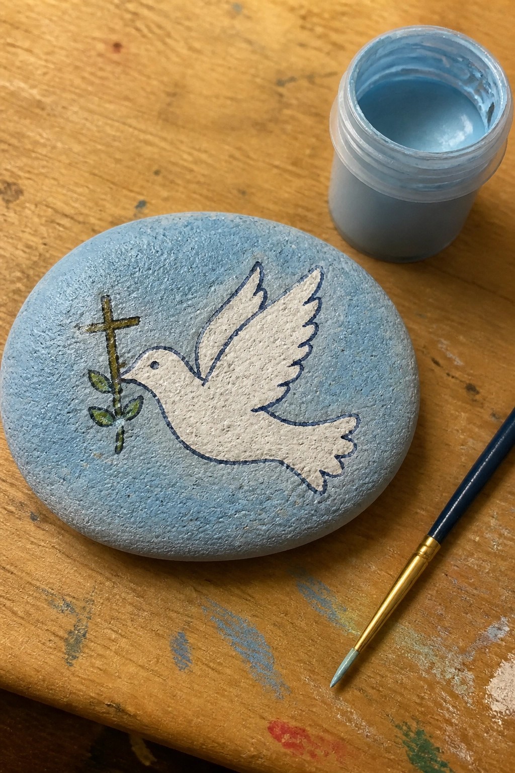 Blue stone with white dove holding cross and olive branch beside paint jar and brush.