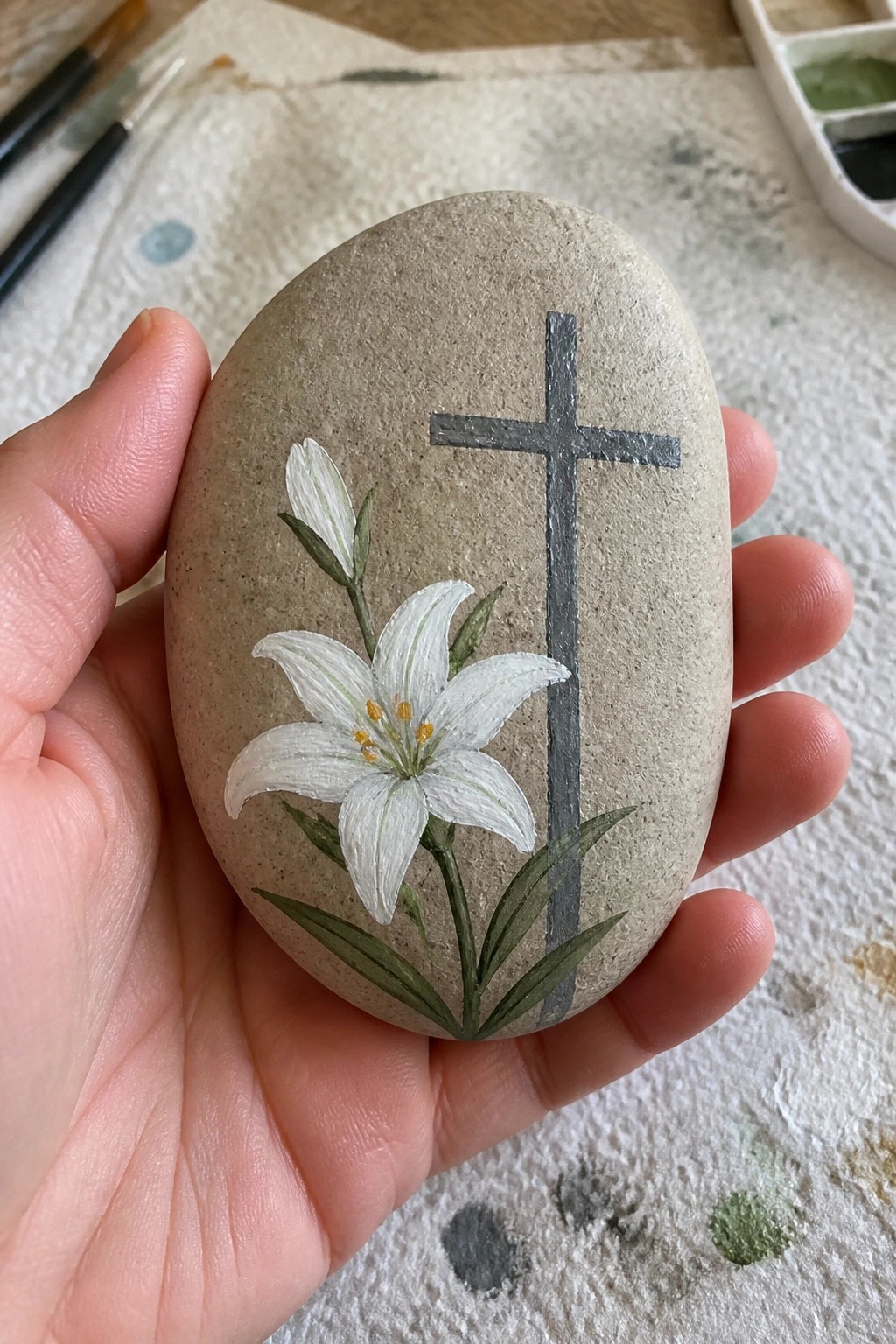 Hand holding smooth stone painted with white lily and gray cross.