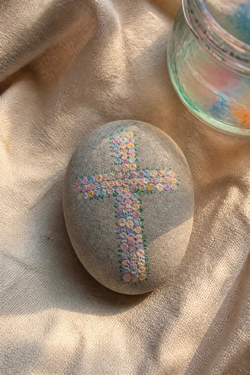 Gray stone with pastel floral cross on beige fabric beside glass jar.