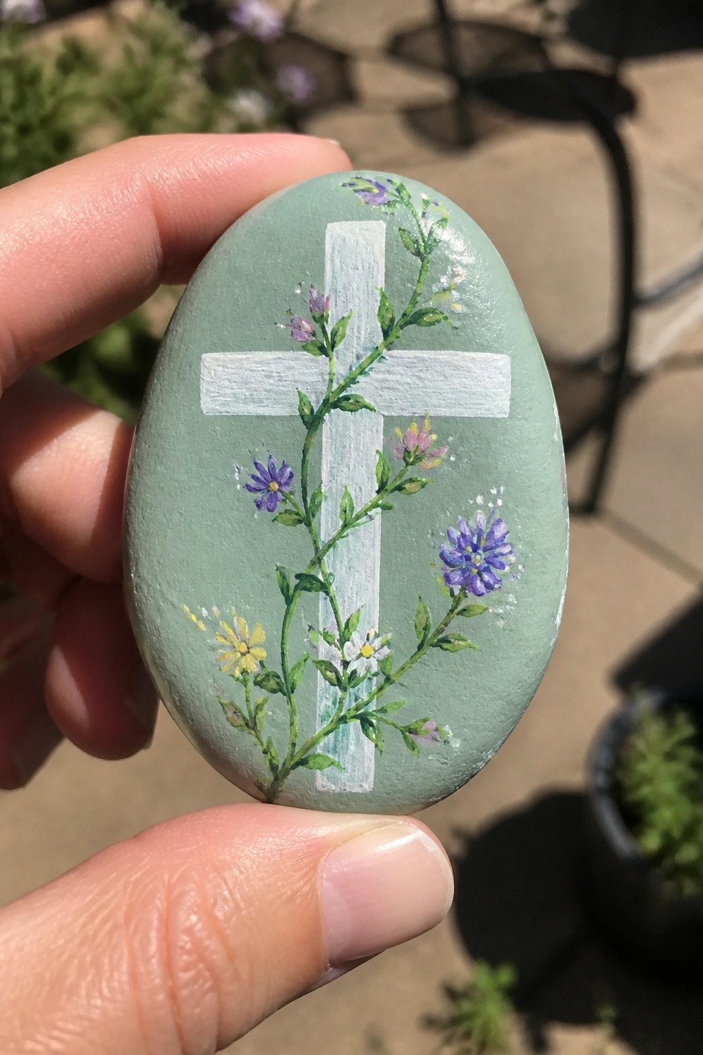 Hand holding a light green painted stone with white cross and floral vines
