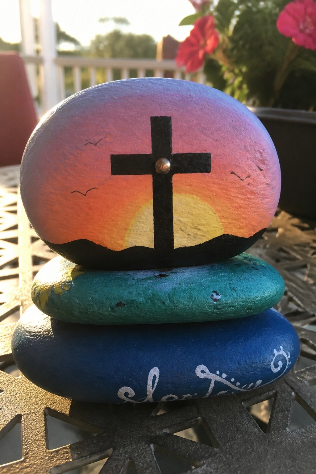 Stacked painted rocks with sunset gradient, black cross, birds, and script text