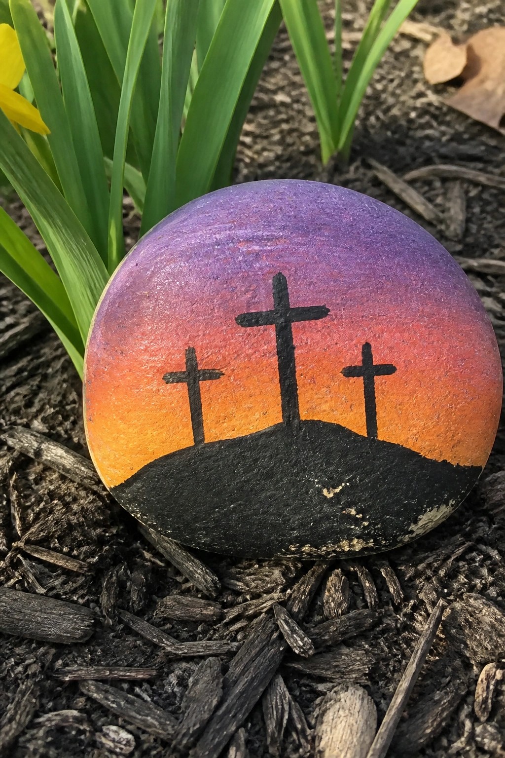 Painted rock with three crosses on a hill under purple-orange sunset sky