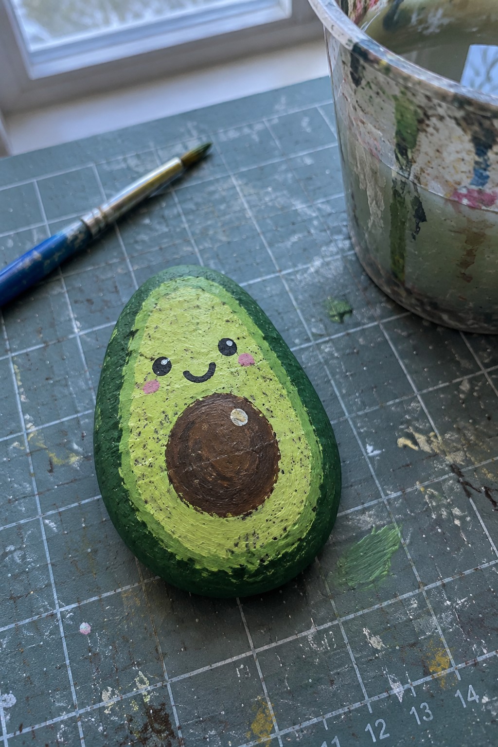 Painted avocado rock with cute face on gridded mat near paintbrush and jar