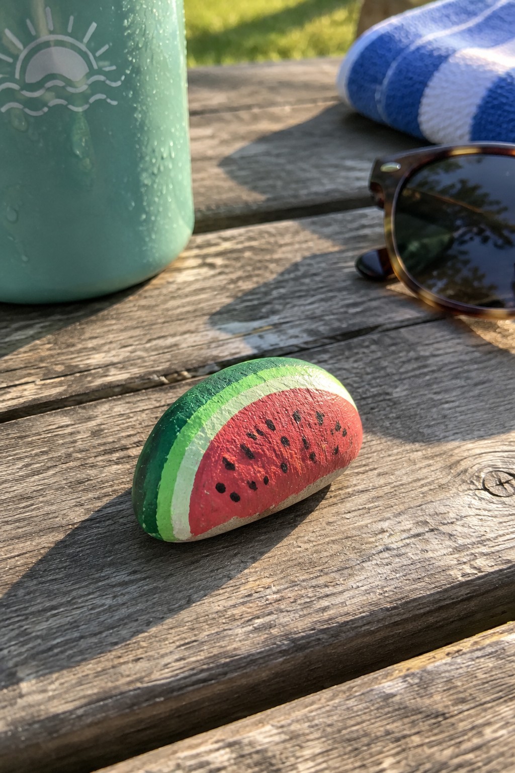 A small rock painted like a watermelon slice sits on a wooden table next to a turquoise water bottle and sunglasses.