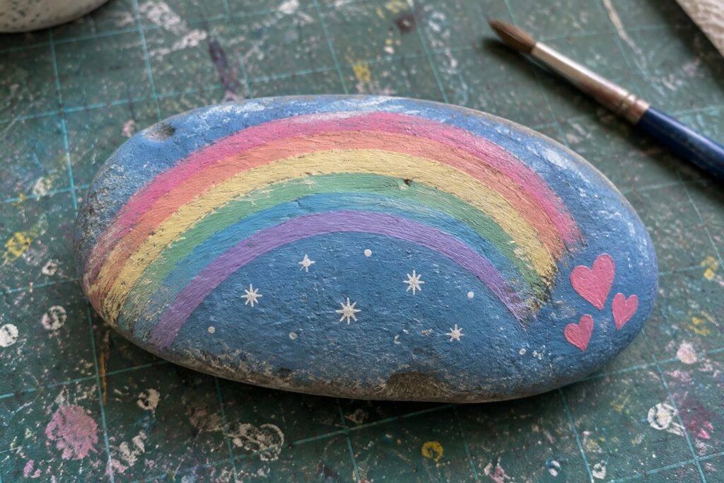 Blue rock with rainbow, stars and hearts on splattered green mat beside paintbrush