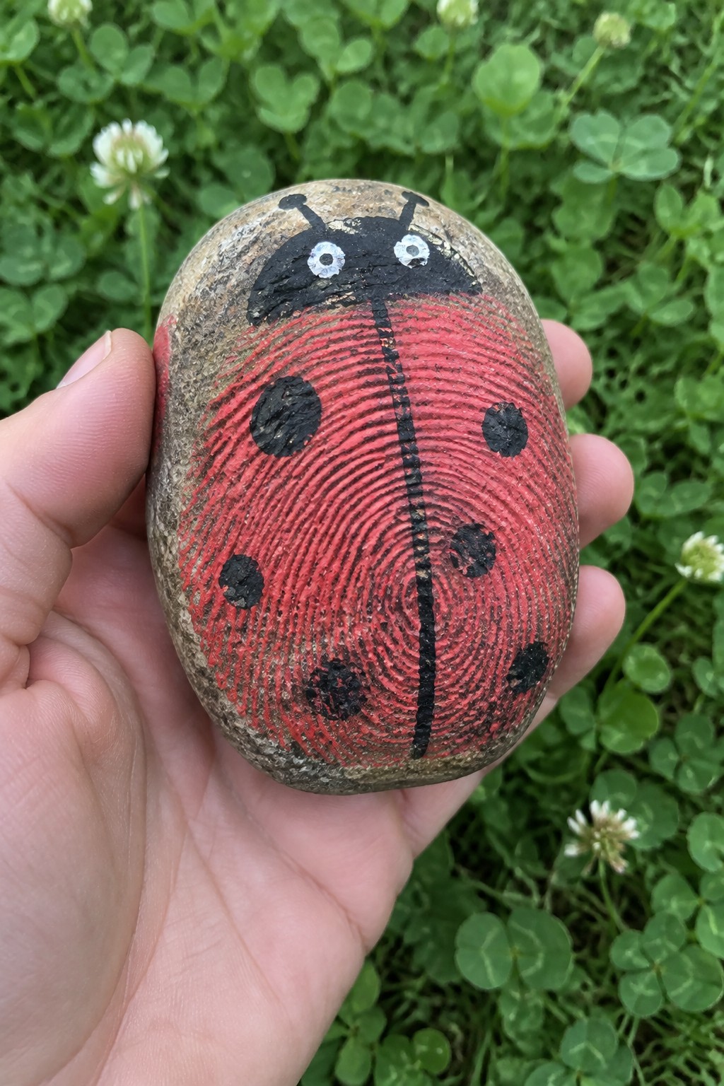 Hand holding red ladybug-painted rock with black spots and fingerprint texture on grass