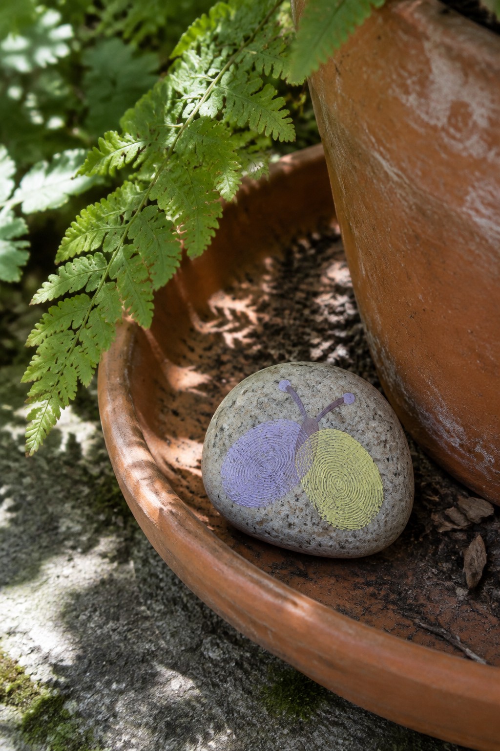 Painted snail on stone with purple and yellow fingerprints in terracotta dish beside fern.