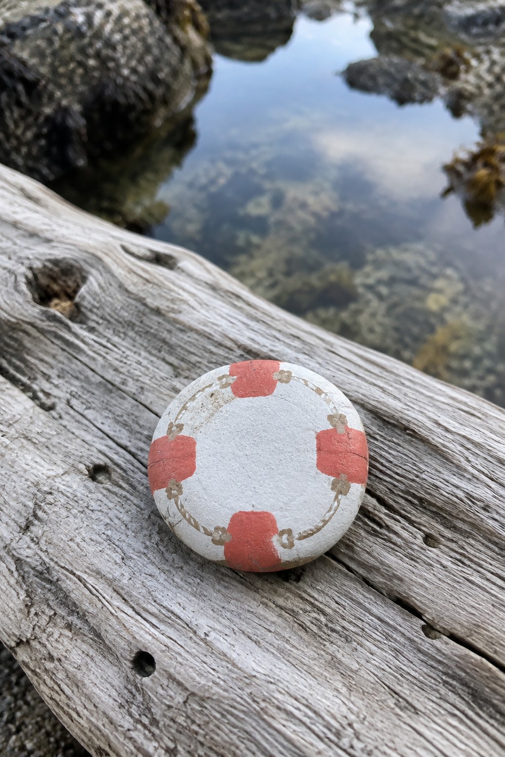 Painted white stone with red lifebuoy design on weathered driftwood by water