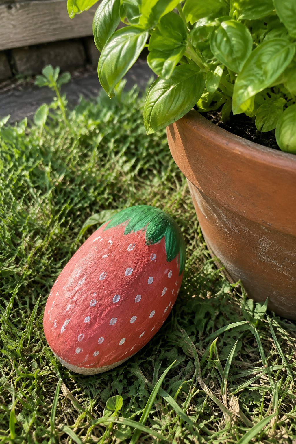 Painted strawberry rock on grass beside terracotta pot with green plant.