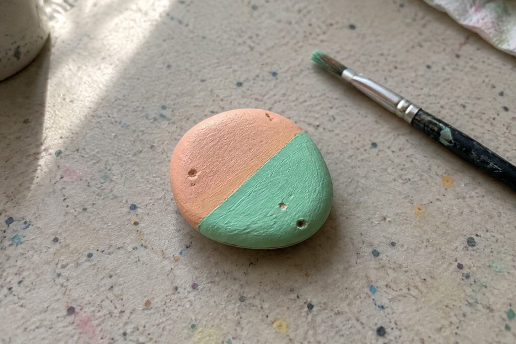 A small rounded rock painted half peach and half mint green sits on a speckled surface next to a paintbrush.