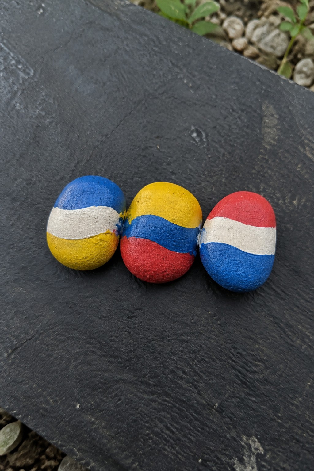 Three painted rocks with blue-white-yellow, yellow-blue-red, red-white-blue stripes on dark slate.