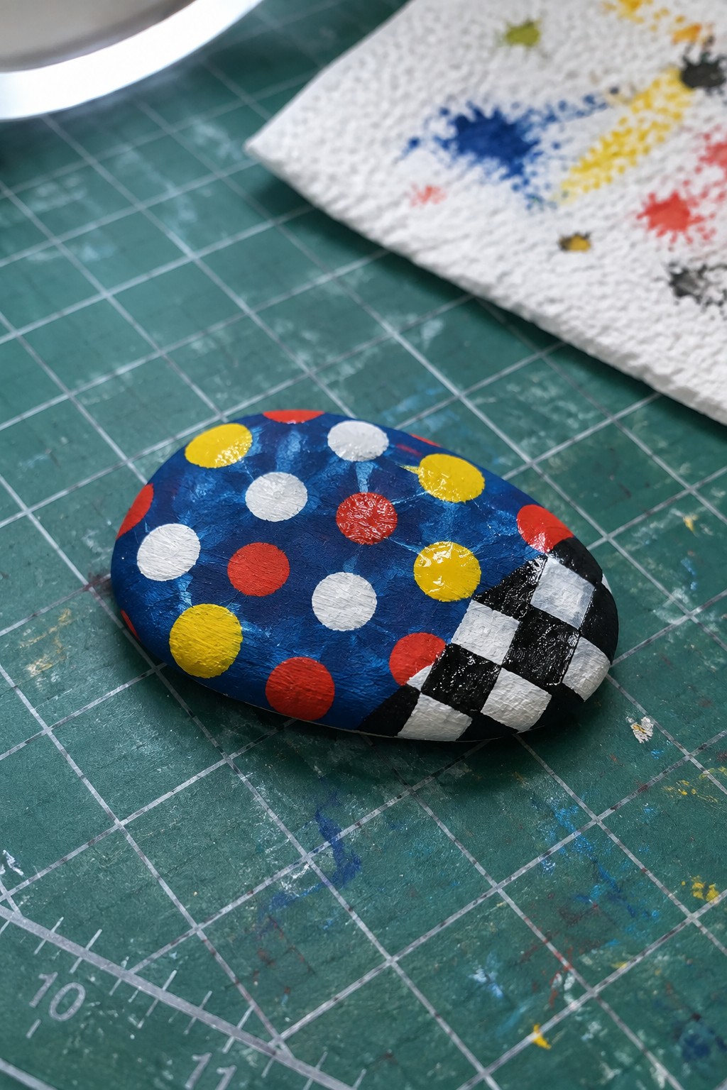 Painted rock with blue background, red white yellow dots, and checkered pattern on green mat