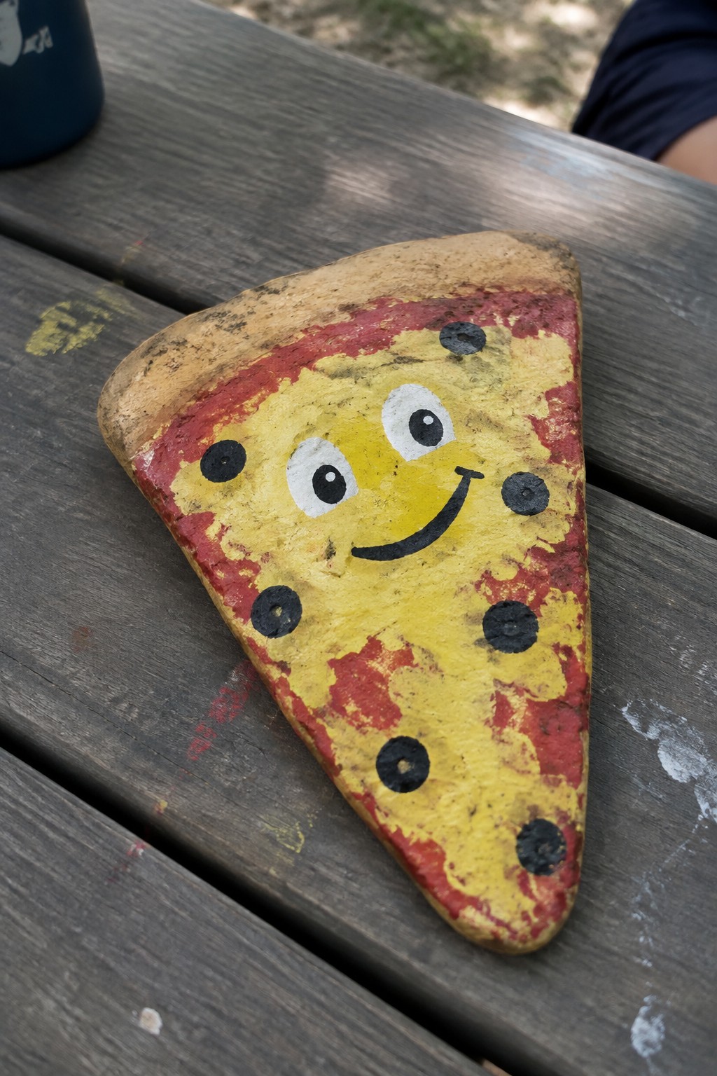Painted rock shaped like smiling pizza slice on weathered wooden picnic table