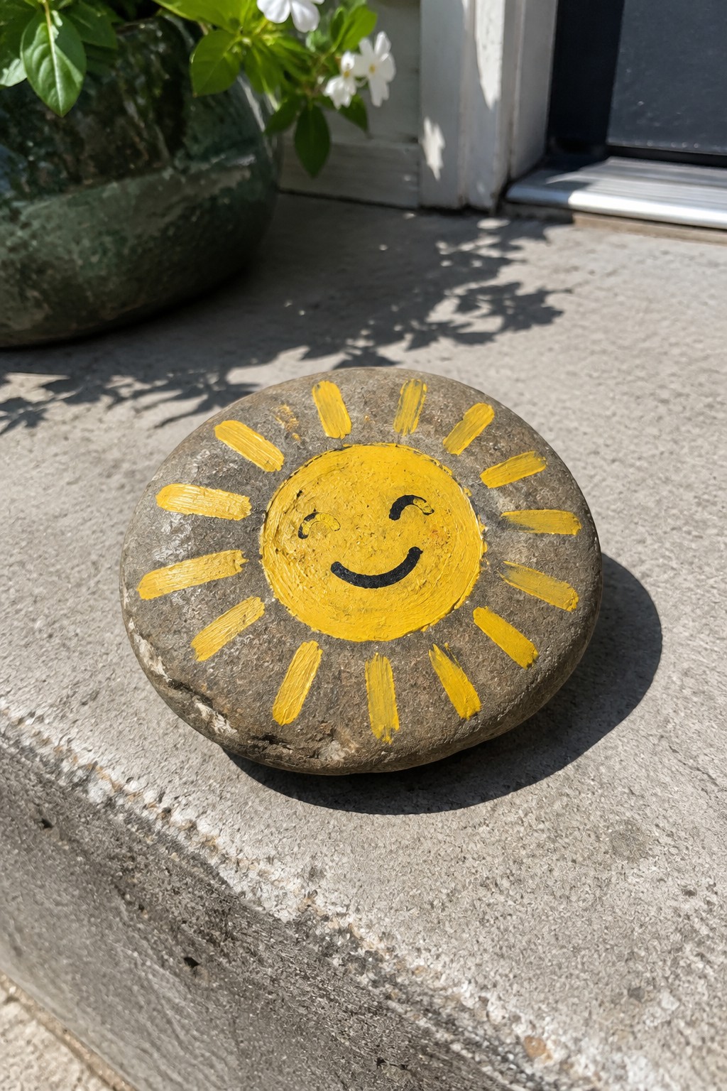 Yellow smiling sun painted on round gray stone on concrete step near plant pot