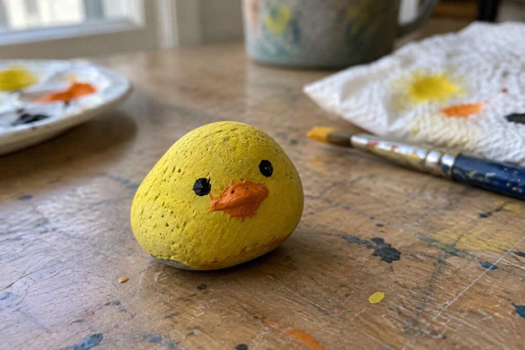 Yellow painted rock chick with black eyes and orange beak on splattered wooden table