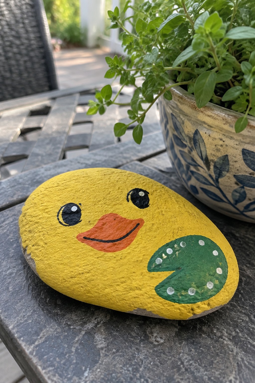 Yellow duck-painted rock with orange beak and green lily pad on table beside plant pot