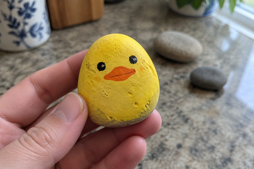 Hand holding yellow painted rock duck with orange beak and black eyes