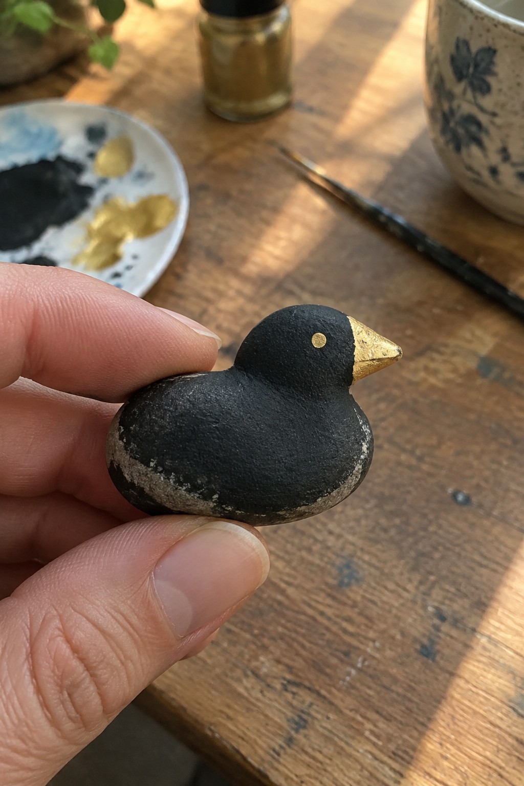 Hand holding small black painted bird with gold beak and eye on wooden table