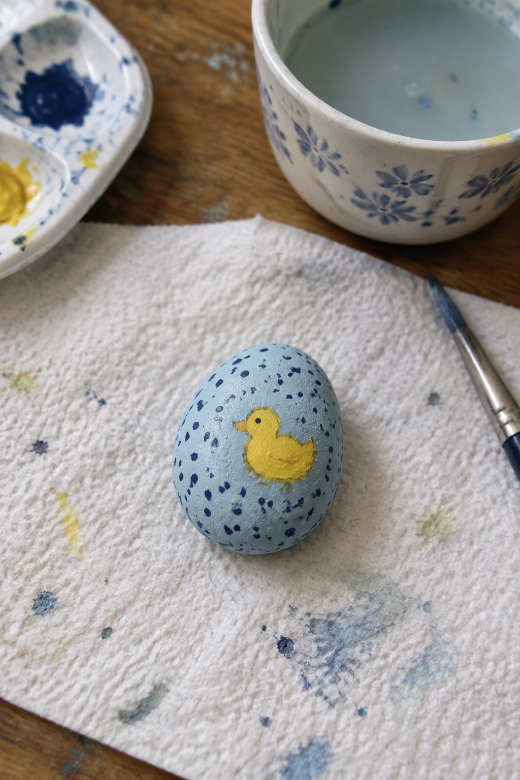 Speckled light blue egg with yellow duckling on paint-stained paper towel beside brush and palette