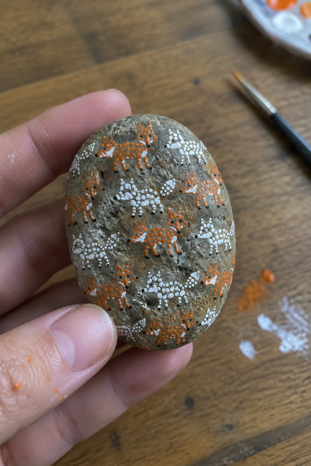 Hand holding oval stone painted with dotted orange and white foxes on wood table