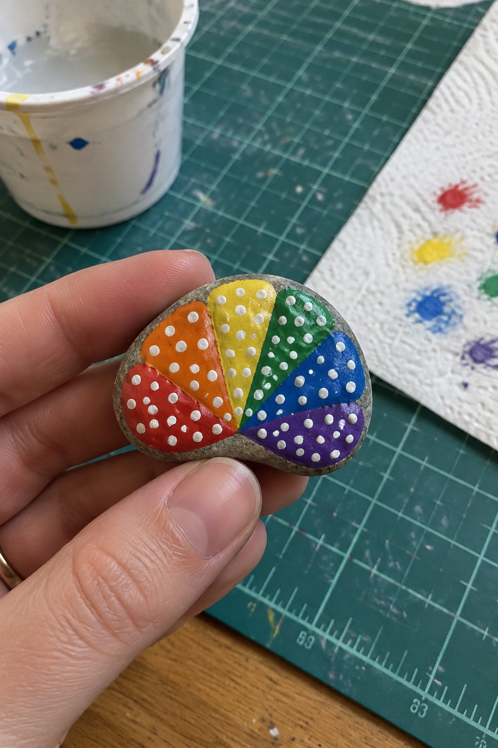 Hand holding rainbow-painted rock with white dots over green cutting mat.