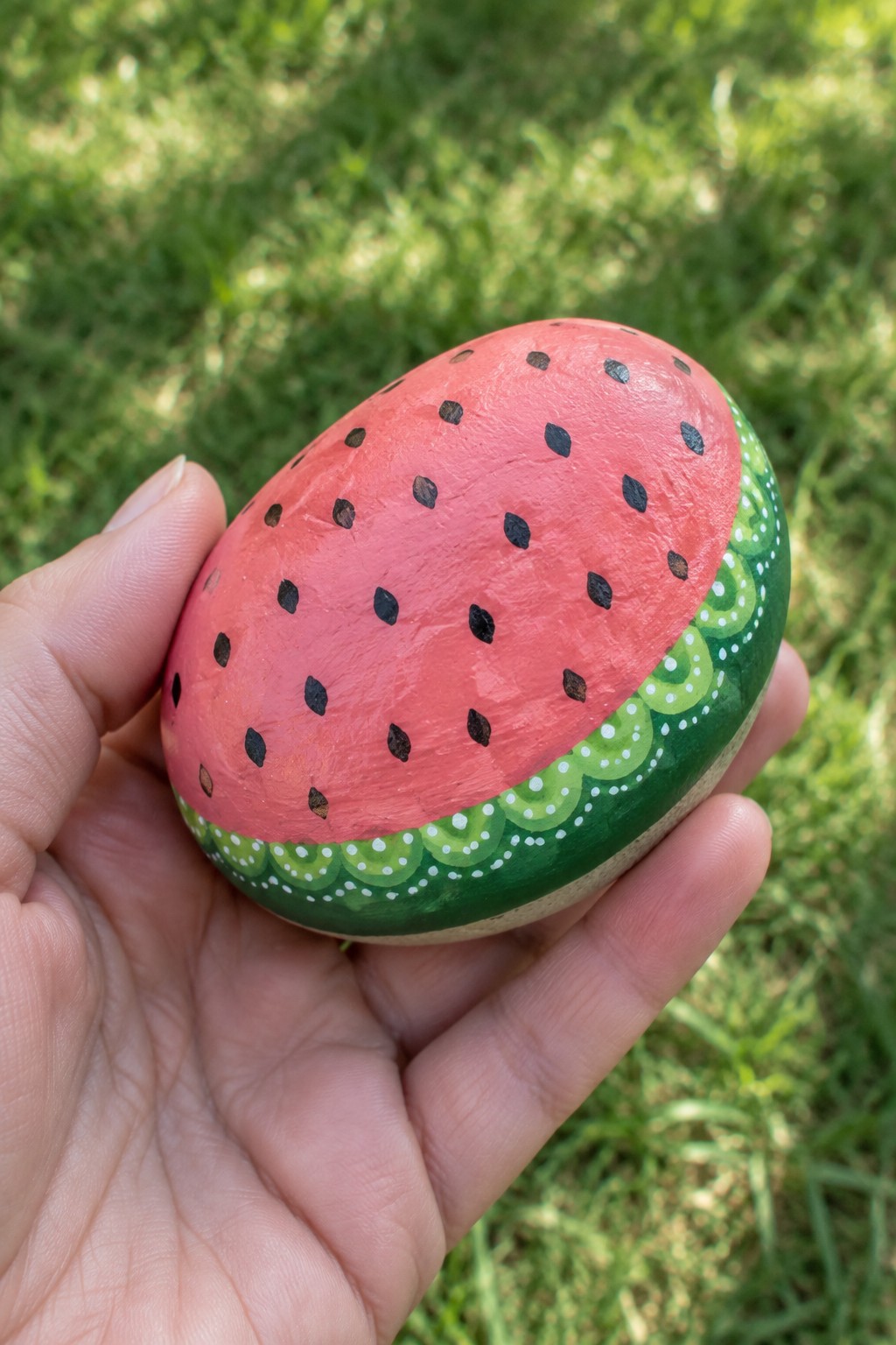 Hand holding oval rock painted red with black seeds and green rind.
