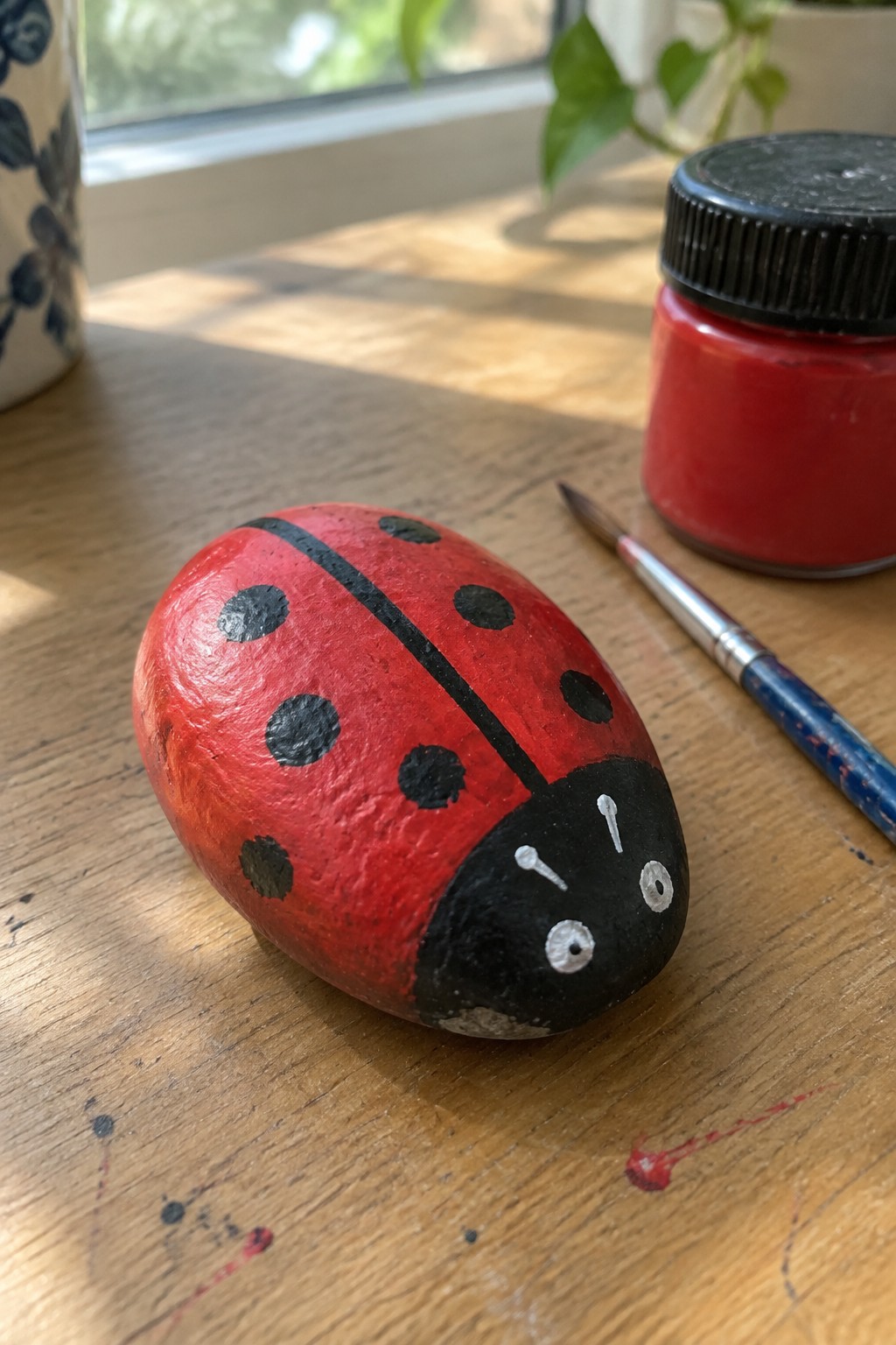 Red ladybug rock painted on wood beside red paint jar and brush