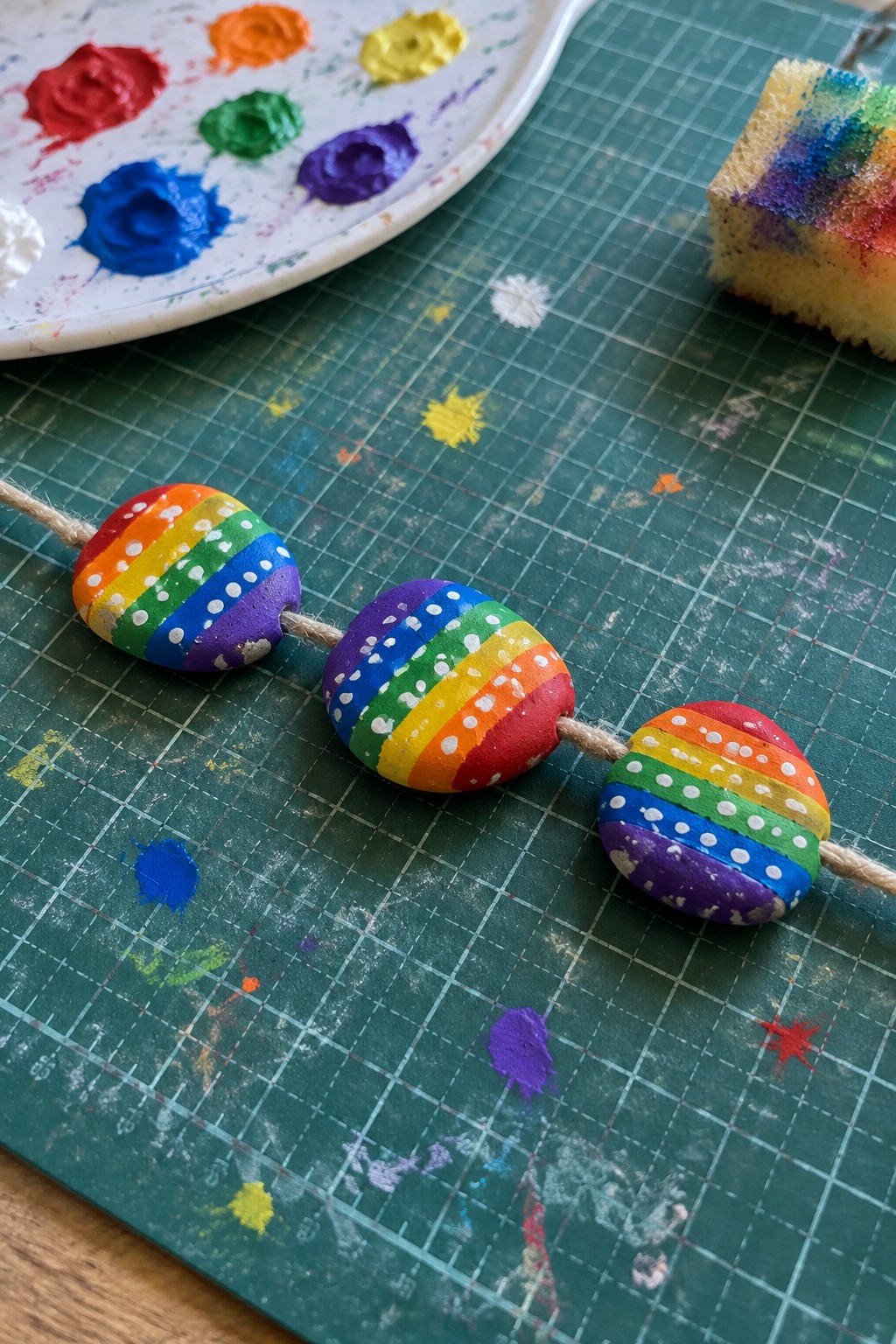 Rainbow painted beads on string beside acrylic paint palette on green cutting mat