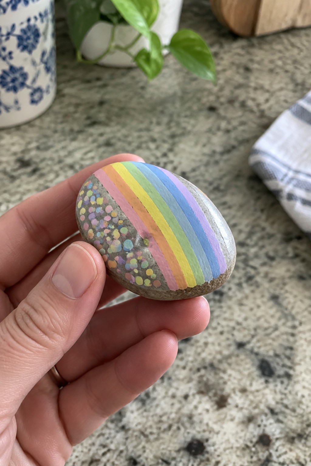 Hand holding oval rock painted with rainbow stripes and colorful dots