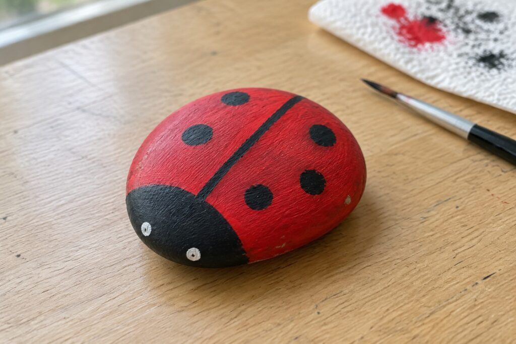 A red and black painted ladybug on a smooth oval rock sitting on a wooden table next to a paintbrush.